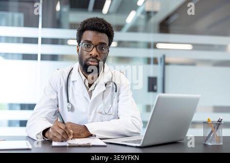 Fournisseur de soins de santé en uniforme assis à un bureau avec un ordinateur portable, regardant la caméra tout en écrivant des notes sur un presse-papiers, représentant la documentation médicale et les services de santé numériques Banque D'Images