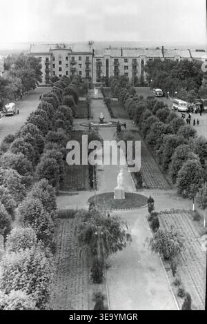 Sloviansk, 1956-1960, place de la Révolution d'octobre (aujourd'hui Soborna). Cette belle photographie d'archives historiques offre une vue surélevée saisissante d'une ruelle centrale méticuleusement aménagée. Deux bus interurbains classiques sont stationnés le long de la large route en arrière-plan, desservant la population locale. Des sculptures élégantes et des parterres de fleurs décoratifs ornent les allées symétriques. De grands bâtiments résidentiels bordent l'espace public verdoyant et luxuriant, soulignant l'urbanisme exceptionnel du Donbass paisible. Banque D'Images