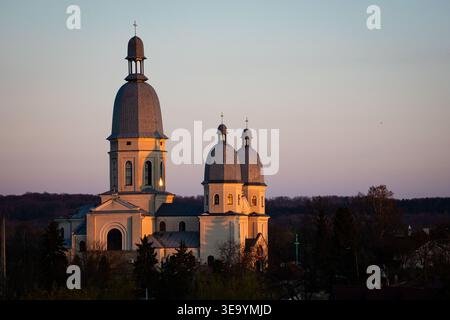 L'élégante église avec un grand clocher et deux dômes brille par la lumière chaude du coucher du soleil au-dessus d'une petite ville. Un ciel pastel doux et une ligne de bois lointaine créent un paix Banque D'Images