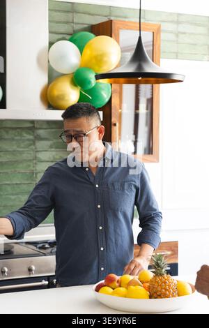 Homme asiatique senior debout au comptoir de la cuisine portant une chemise bleue et des verres arrangeant un bol de fruits Banque D'Images