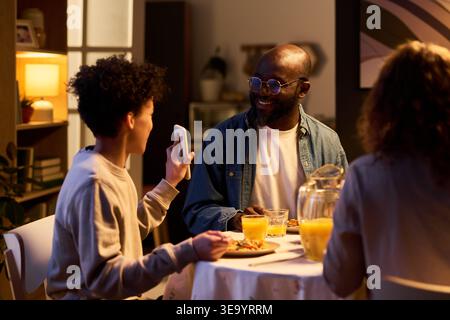 Père afro-américain souriant à un adolescent montrant son smartphone pendant le dîner de famille, discutant du contenu numérique et de la connexion. Idéal pour les parents, le temps d'écran, le marketing de style de vie familial Banque D'Images