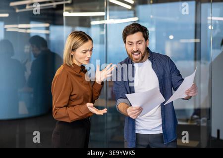 Deux collègues d'affaires frustrés se disputent dans un bureau moderne : un homme en colère criant tout en saisissant des documents et une femme confuse faisant des gestes en désaccord lors d'une réunion tendue Banque D'Images