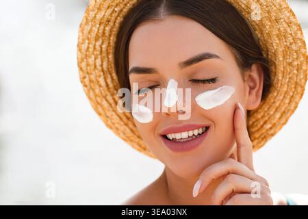 Femme portant un chapeau de paille appliquant de la crème solaire sur la joue. Jeune femme souriant à la lumière du soleil. Modèle féminin touchant doucement le visage. Soins de la peau et protection solaire c Banque D'Images