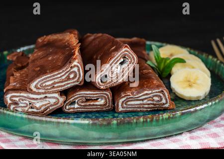Délicieux rouleaux de crêpe au chocolat remplis de crème servis sur une assiette avec des tranches de bananes fraîches, gros plan. Concept de dessert parfait pour la nourriture de menu de café pho Banque D'Images