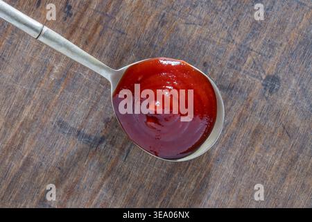 Ketchup tomate rouge ou sauce BBQ à la cuillère sur bois rustique. Gros plan de sauce rouge épaisse dans une cuillère en métal sur fond de bois altéré. Parfait pour Cu Banque D'Images