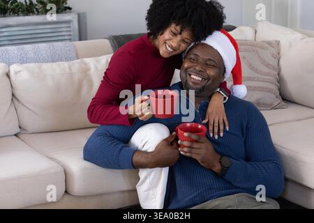 Couple afro-américain assis sur le canapé en coupe beige, homme portant le chapeau de Père Noël et tenant des tasses rouges. Cosy, festif, chaleureux, affection, ensemble, souriez, à la maison Banque D'Images