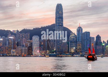 Jonque traditionnelle en bois avec des voiles rouges contre Hong Kong skyline pendant le coucher du soleil, scène urbaine emblématique Banque D'Images