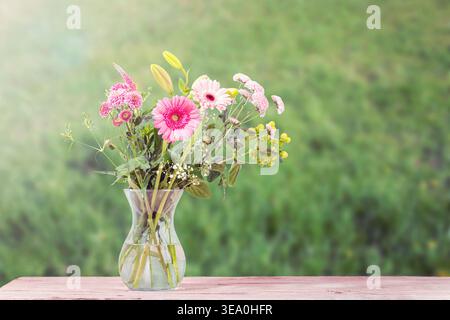 Bouquet coloré de fleurs fraîches dans un vase en verre sur une table en bois avec un fond flou vert Banque D'Images