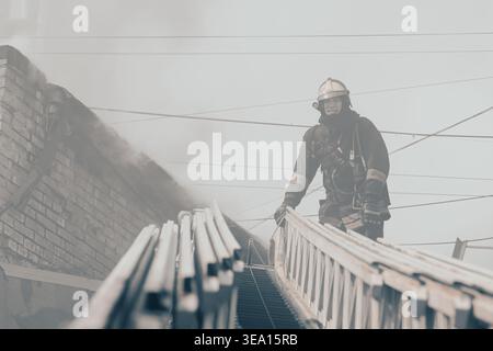 Krasnoïarsk, Russie, 14 04 2026 : pompier grimpant une échelle pendant une urgence incendie dans un bâtiment. Une fumée épaisse entoure la scène comme opérat de sauvetage Banque D'Images