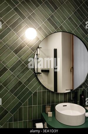 Intérieur moderne de la salle de bains avec carreaux vert émeraude en diagonale, miroir circulaire, évier blanc et luminaires noirs mats. Banque D'Images