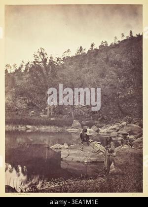 Vue sur Apache Lake, Sierra Blanca Range, Arizona, deux scouts Apache au premier plan par Timothy O'Sullivan, 1873, montrant des scouts indigènes positionnés sur la rive rocheuse à côté de l'eau calme et de la forêt dense Banque D'Images