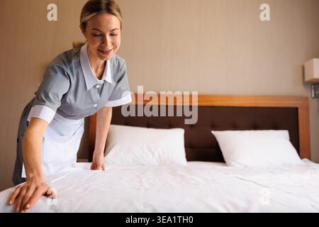 Femme de ménage professionnelle de l'hôtel faisant lit avec draps blancs frais dans l'intérieur moderne de la chambre Banque D'Images