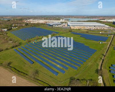 Vue aérienne de panneaux solaires disposés en rangées sur un champ vert près d'entrepôts industriels sous un ciel bleu à Coalville, Angleterre, Royaume-Uni. Banque D'Images