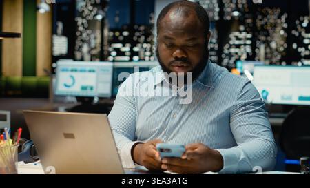Employé de bureau noir vérifiant une notification connectée sur les réseaux sociaux pendant qu'il fait des heures supplémentaires à minuit, vérifiant smartphone au travail après les heures de travail. Jeune homme lisant des messages texte. Banque D'Images