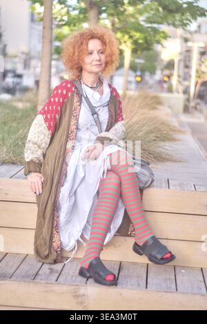 Portrait d'une femme gingembre d'âge moyen aux cheveux bouclés portant une tenue excentrique avec des collants rayés et un béret bleu. Elle est assise sur un bois Banque D'Images