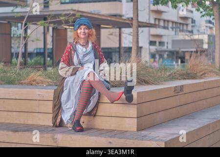 Portrait d'une femme gingembre d'âge moyen aux cheveux bouclés portant une tenue excentrique avec des collants rayés et un béret bleu. Elle est assise sur un bois Banque D'Images
