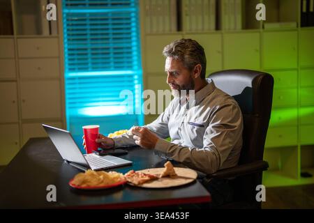 Bureau de nuit concentré avec ordinateur portable et collation. Détente au bureau de nuit. Travail de nuit. Homme d'affaires manger collation tout en travaillant sur ordinateur portable. Homme d'affaires Banque D'Images