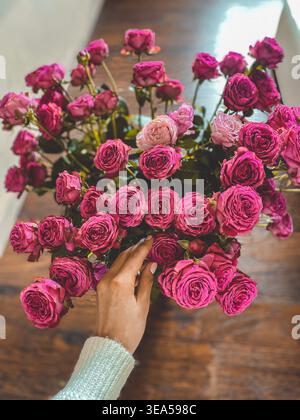 Bouquet de roses roses avec une main féminine touchant des fleurs sur une table en bois, lumière naturelle douce et atmosphère romantique. Banque D'Images