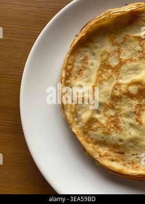 Pile de fines crêpes sur une assiette blanche posée sur une table en bois, composition minimale avec lumière naturelle. Banque D'Images
