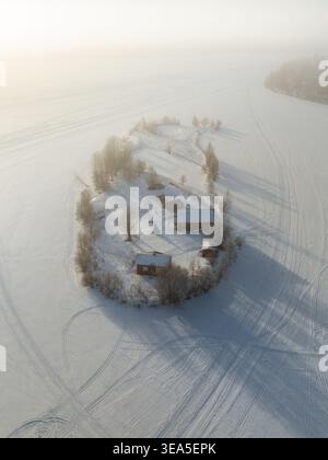 Vue aérienne d'une petite île avec des cabanes traditionnelles en bois et des arbres enneigés entourés d'un lac gelé dans un paysage hivernal brumeux à Rovanie Banque D'Images
