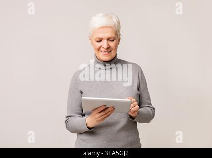 Femme senior avec les cheveux blancs courts en utilisant tablette et souriant dans un cadre neutre Banque D'Images