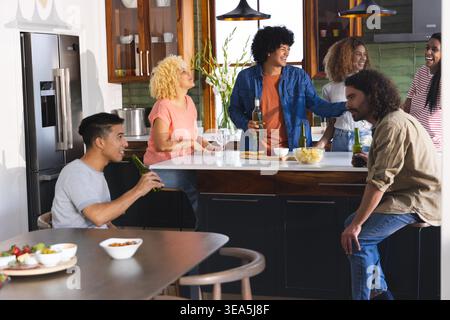 Divers amis discutant et tenant des boissons dans la cuisine moderne autour de l'île blanche avec des frites. Convivial, rassemblement, décontracté, boissons, comptoir, suspension, éclairage Banque D'Images