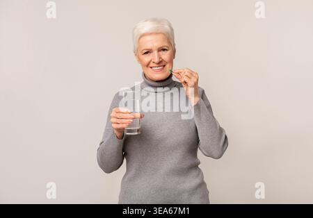Femme senior avec les cheveux blancs courts prenant un supplément avec de l'eau dans un fond plat Banque D'Images