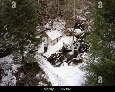 Vue aérienne du moulin en pierre du Moulin de Chambeuil niché dans une forêt enneigée à côté d'une cascade et d'un ruisseau à Laveissière, Auvergne-Rhône-Alp Banque D'Images