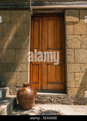 Double porte en bois brun encadrée par des murs en pierre taillée rugueuse. Un grand pot décoratif en argile repose sur des marches en pierre ensoleillée menant à l'entrée. Banque D'Images