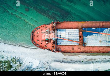 Une vue aérienne montre un grand cargo rouge abandonné échoué sur une côte de galets blancs. Banque D'Images