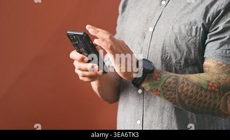Homme tatoué à l'aide d'un smartphone avec Smartwatch dans une chemise décontractée - gros plan des mains texturant Banque D'Images