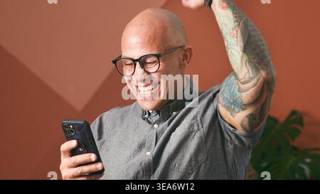 Homme d'affaires tatoué chauve célébrant avec smartphone dans le bureau moderne - victoire heureuse de l'entrepreneur Banque D'Images