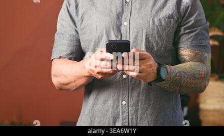 Homme tatoué à l'aide d'un smartphone dans une chemise décontractée montrant des mains, montre et mode de vie de communication mobile Banque D'Images