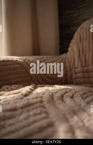 gros plan ensoleillé d'un canapé beige en peluche drapé d'un jet texturé dans un salon confortable, tons neutres chauds, toile de fond de rideau doux et motif subtil Banque D'Images