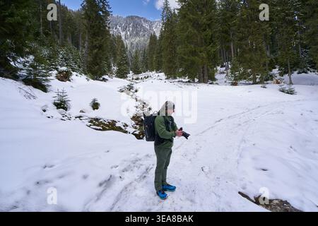 Un homme mûr examine son appareil photo dans une forêt couverte de neige avec un ruisseau de fonte des neiges et un pic de montagne rocheux s'élevant à travers les épinettes Behi Banque D'Images