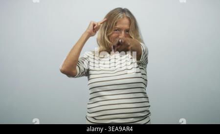Femme blonde senior pointe le doigt directement sur la caméra tout en faisant des gestes vers sa tempe, portant une chemise rayée dans le studio ; défi affirmé. Banque D'Images