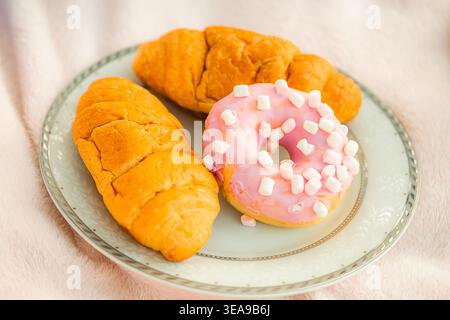 Deux croissants et un beignet reposent sur une assiette bleue avec un bord doré sur une serviette blanche. Pique-niquez pendant les vacances d'été et les loisirs en plein air. Banque D'Images