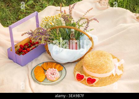 Deux croissants et un beignet reposent sur une plaque bleue avec un rebord doré sur une couverture blanche. À côté se trouve un chapeau de paille et des verres en forme de coeur, panier avec daisi Banque D'Images