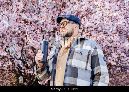 Joyeux homme barbu tenant la coupe Thermos près de Cherry Blossoms Banque D'Images