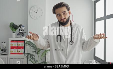 Homme avec stéthoscope haussant les épaules avec les mains dans un bâtiment de bureau médical blanc, portant un blouse de laboratoire et la barbe visible ; dilemme de soin de l'incertitude. Banque D'Images