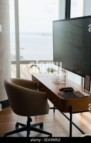 Bureau en bois pour ordinateur portable et téléviseur grand écran. Petite plante verte se trouve à côté de la bouteille d'eau. Horizon de la ville visible à travers de hautes fenêtres. Chaise ergonomique f Banque D'Images