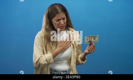 Femme tient le billet de banque en dollar américain entre les doigts et appuie main à poitrine tout en grimacant dans le fond bleu studio ; perte regret tristesse. Banque D'Images