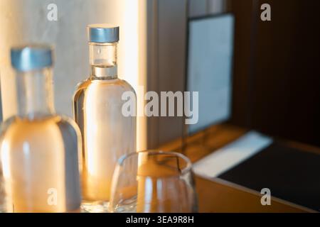Deux bouteilles en verre transparent avec des bouchons argentés reposent sur une table en bois. Un verre à boire vide se tient à proximité. Un éclairage doux crée une lueur chaleureuse. Ordinateur portable scre Banque D'Images