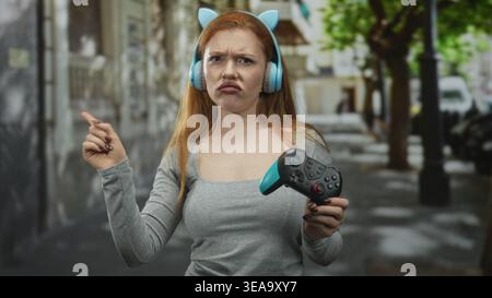 Femme rousse tenant une manette de jeu et portant des écouteurs d'oreille de chat pointe le doigt dans une rue ; défiance de frustration. Banque D'Images