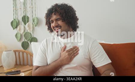 Jeune homme indien serrant la poitrine et appuyant main contre poitrine sur le lit dans le studio avec table de chevet et smartphone, suggérant heartattack ; douleur. Banque D'Images