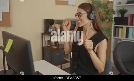 Femme écoutant avec des écouteurs, souriant et tapotant les doigts à l'ordinateur tout en portant des lunettes et gilet noir au bureau ; bonne concentration. Banque D'Images