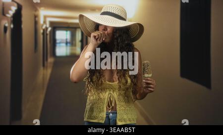 Femme adolescente hispanique tient une barre de crème glacée saupoudrée et presse la main contre la poitrine tout en grignant dans un couloir de bâtiment ; préoccupation de nausées. Banque D'Images