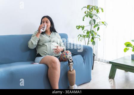 Femme enceinte avec une jambe prothétique hydratant tout en étant assise confortablement sur un canapé à la maison Banque D'Images