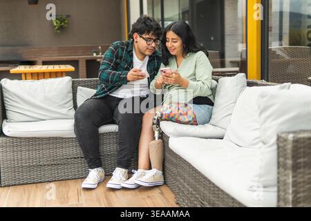 Jeune couple diversifié regardant des smartphones, femme montrant une jambe prothétique, profitant du temps ensemble sur un patio Banque D'Images