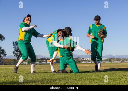 Équipe masculine de baseball diversifiée en uniformes verts sur le terrain célébrant à genoux et serrant avec des gants Banque D'Images
